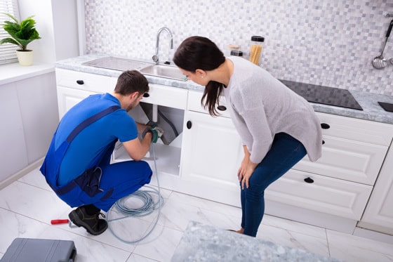 A plumber working under a sink with a customer watching what he's doing.